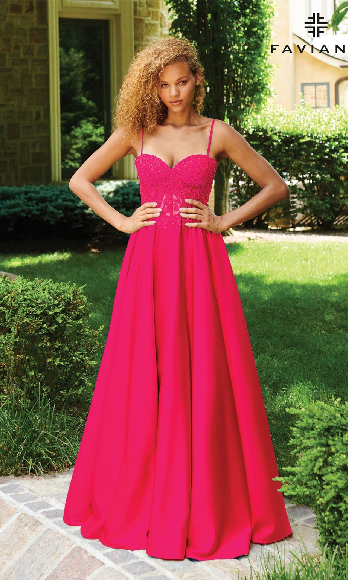 A person stands outside on a stone path wearing the Faviana 11259 Lace-Up Long A-Line Prom Dress in bright pink, featuring thin straps. Greenery and part of a brick building appear in the background.